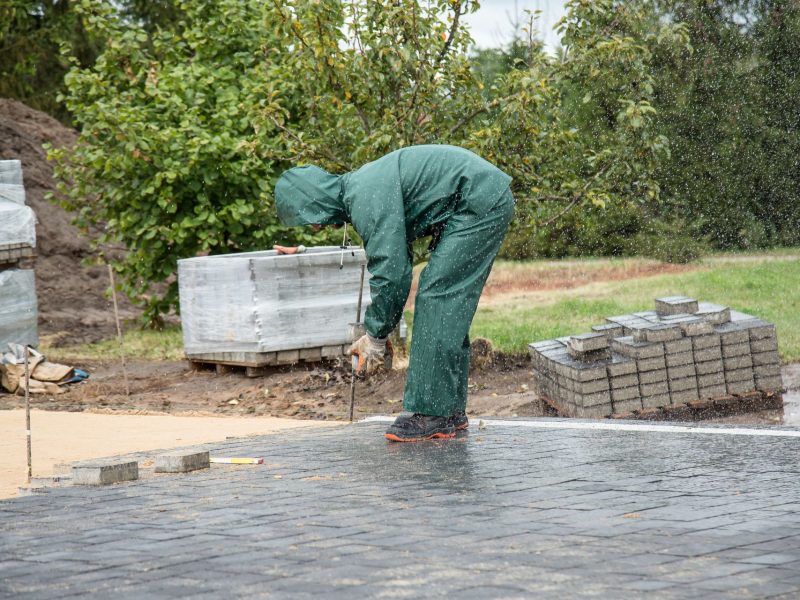 Arbeiter in Regenkleidung verlegt Pflastersteine auf einer Gartenfläche